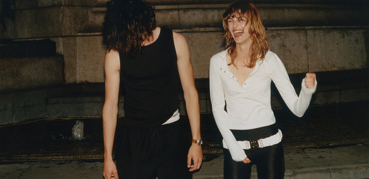 Two people laughing outside at night in front of a stone wall, one in a black tank top, the other in a white long-sleeve top and black leggings